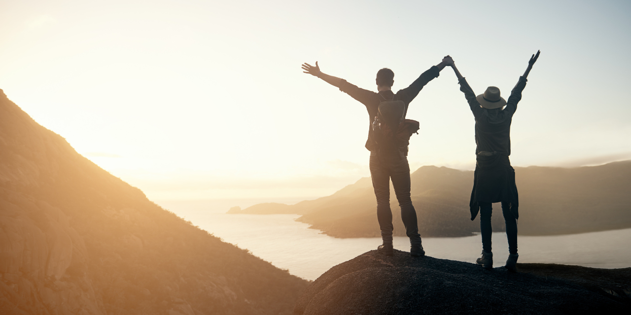 Two people standing on top of a mountain looking out into the distance with their hands in the air.