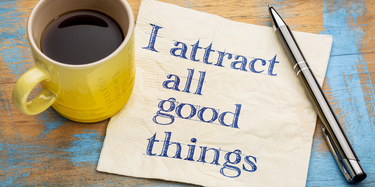 A table with a piece of paper on it that says "I attract all good things" with a pen to the right, and a yellow coffee cup to the left.