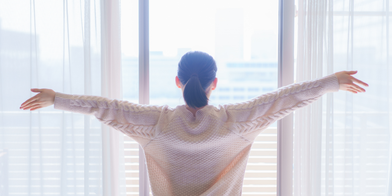 Person standing with arms wide open while looking outside at the sunny day.