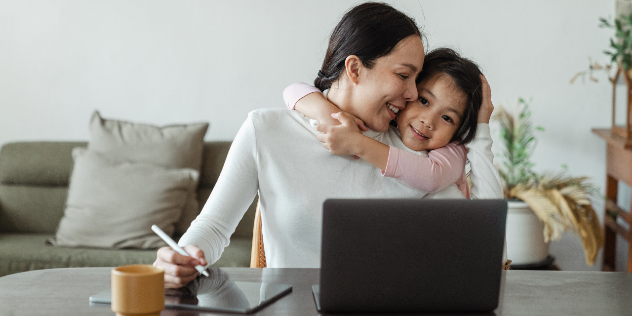 A mother working on her computer, while looking over at her daughter smiling at her while her daughter hugs her.